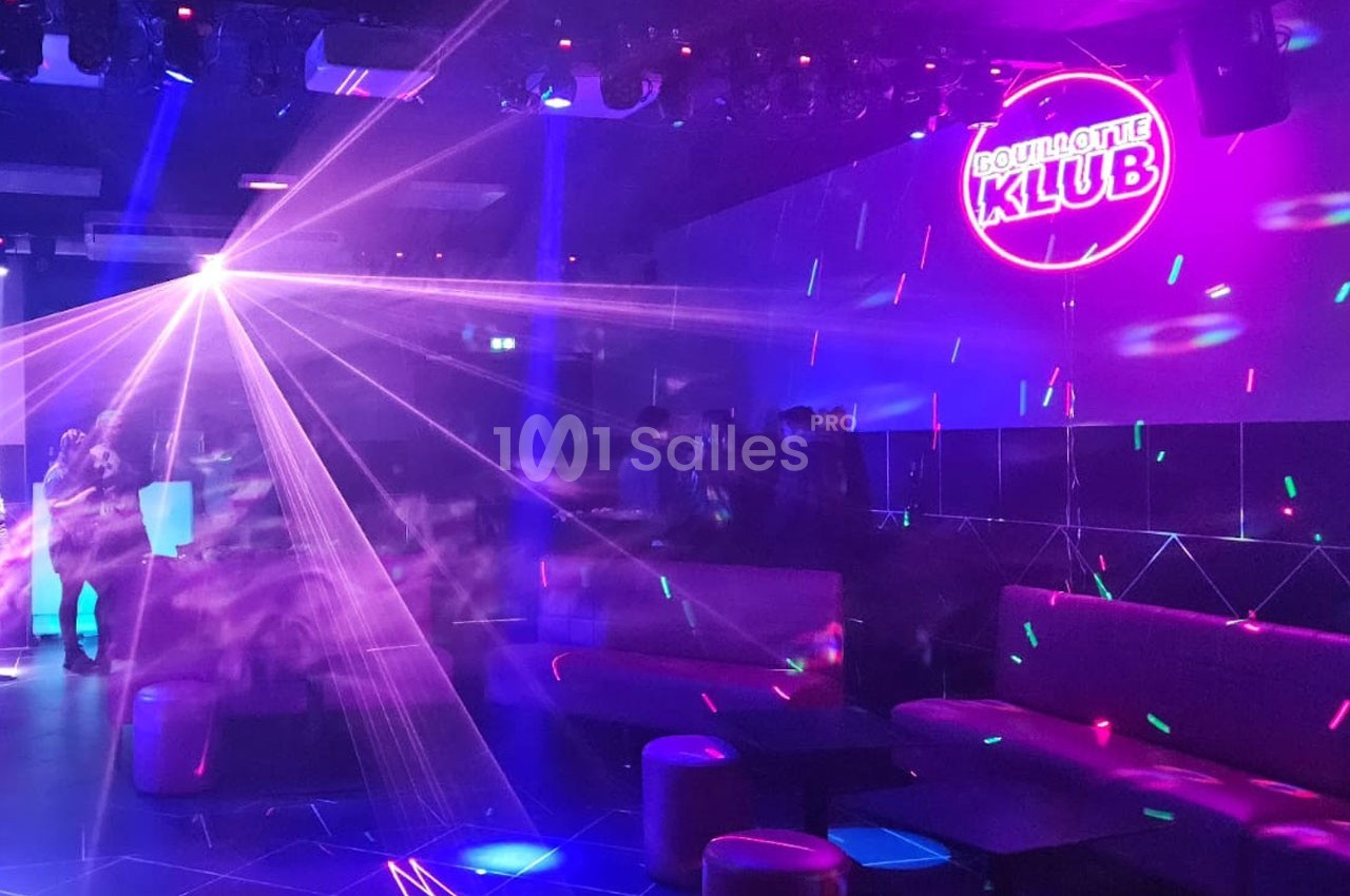 Salle de discothèque avec jeux de lumières colorées, néons et quelques personnes autour de tables basses.