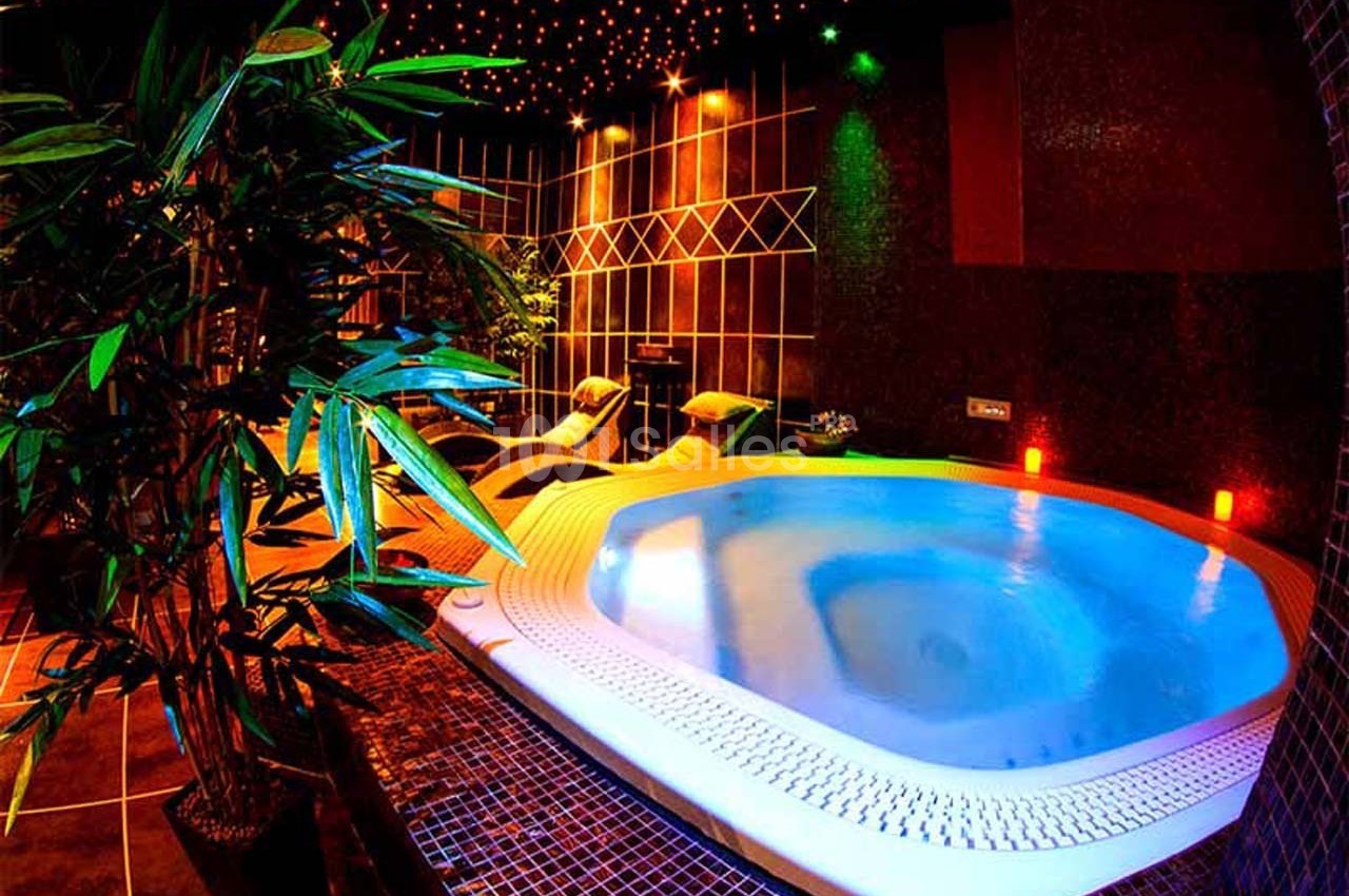 Bain à remous éclairé par des lumières colorées, entouré de plantes et de chaises longues dans une ambiance tamisée.