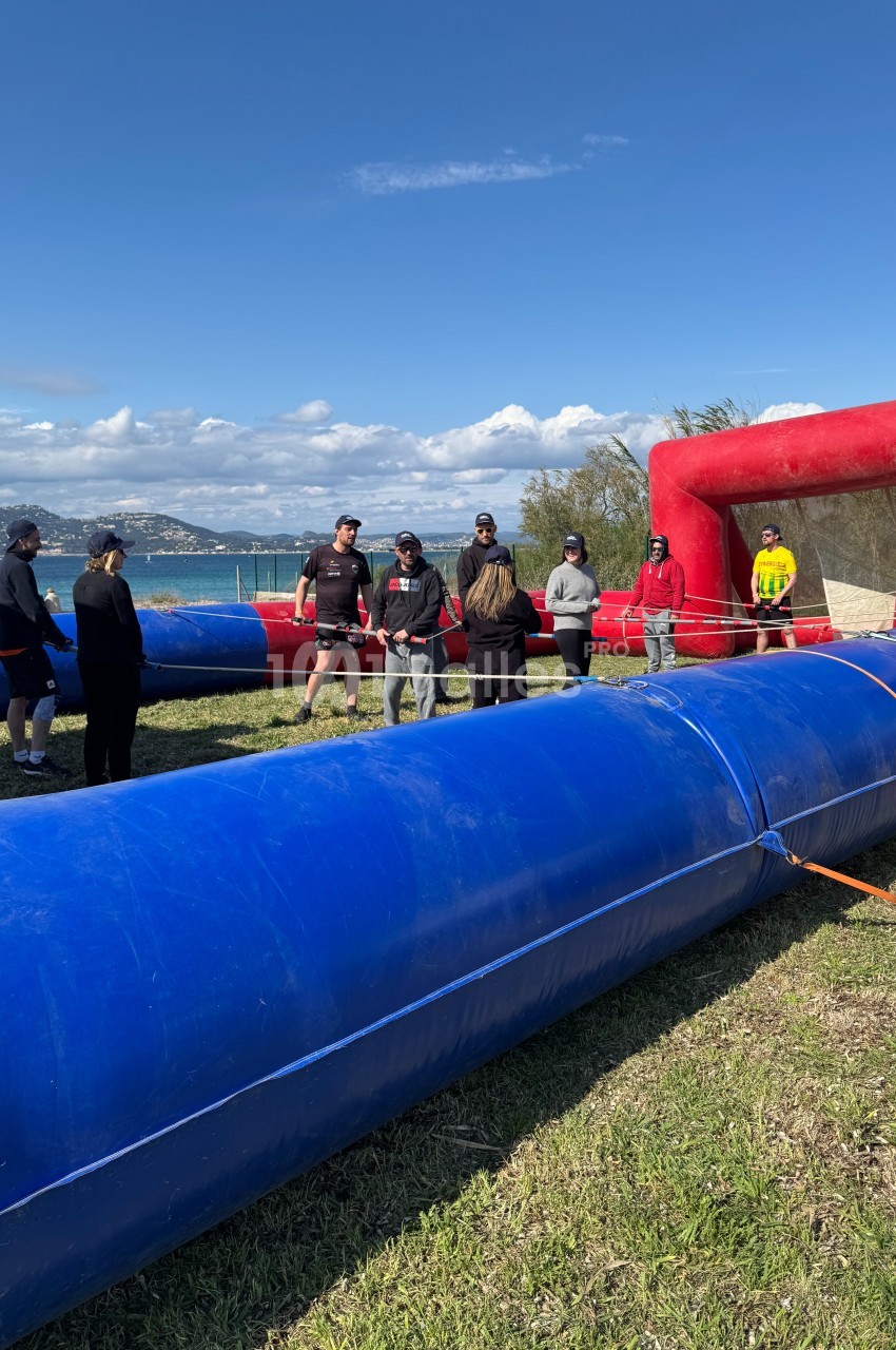 Des personnes debout autour d'un terrain gonflable bleu et rouge en plein air, près d'un plan d'eau.