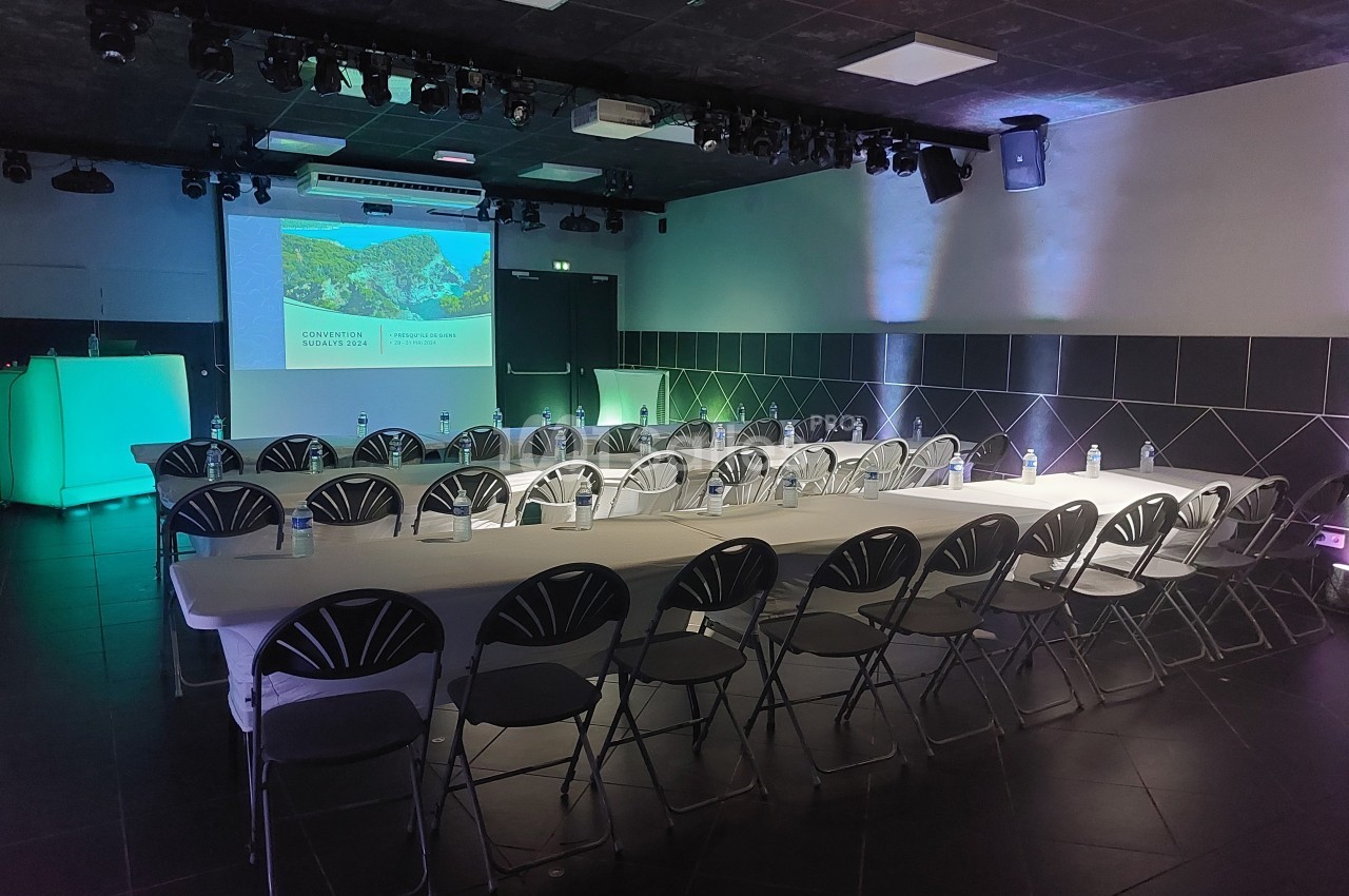 Salle de conférence sombre avec des rangées de chaises et une table, éclairée par des lumières colorées et un projecteur.