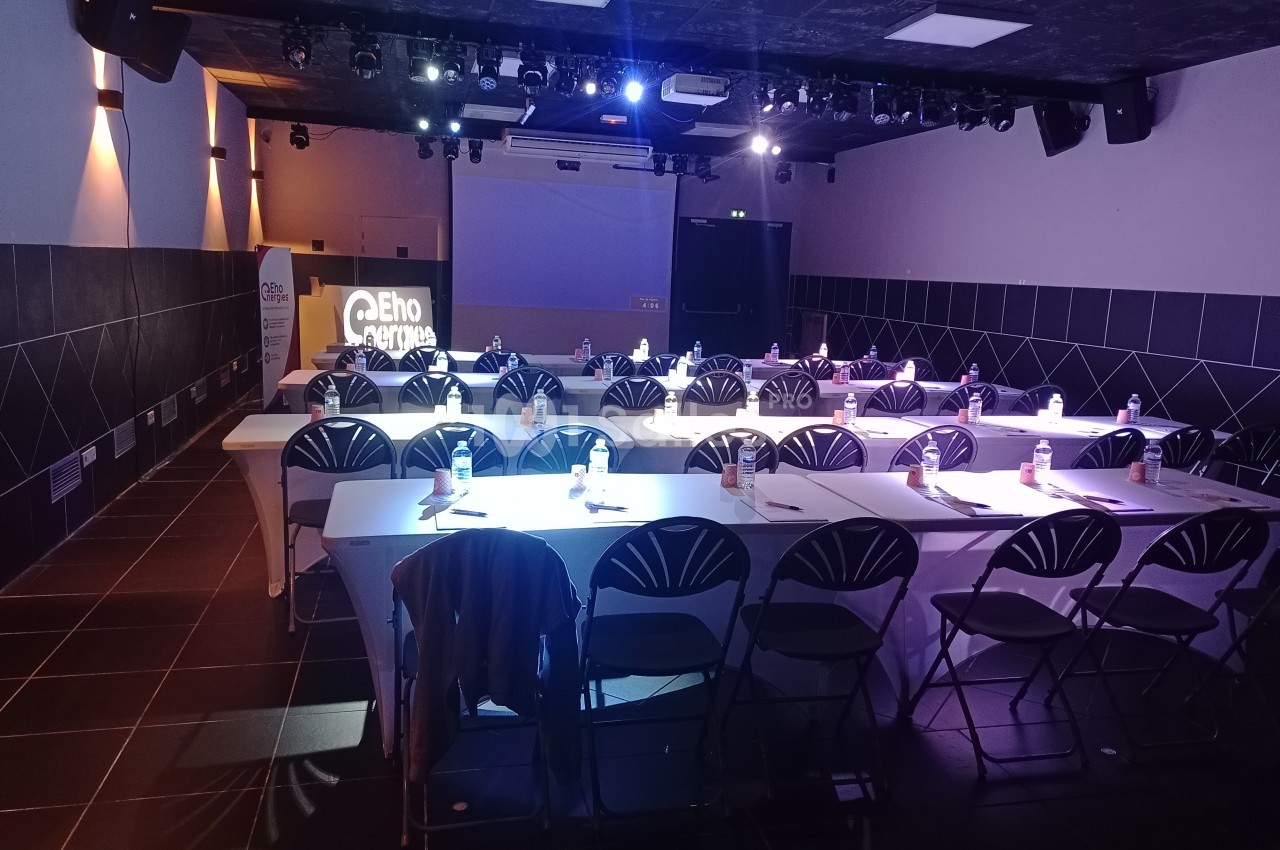 Salle de conférence sombre avec des tables alignées, chaises pliantes et projecteur affichant un logo sur un écran.