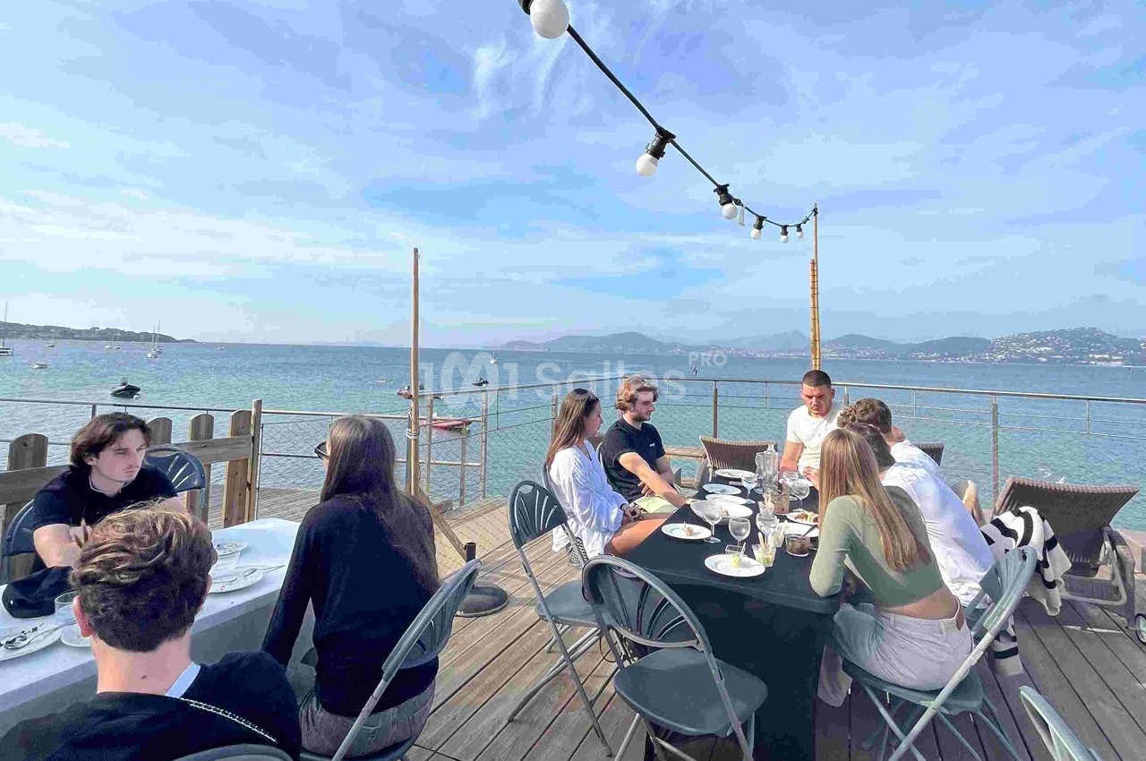 Des personnes dînent en terrasse en bord de mer, avec une vue sur l'eau et les montagnes à l'horizon.