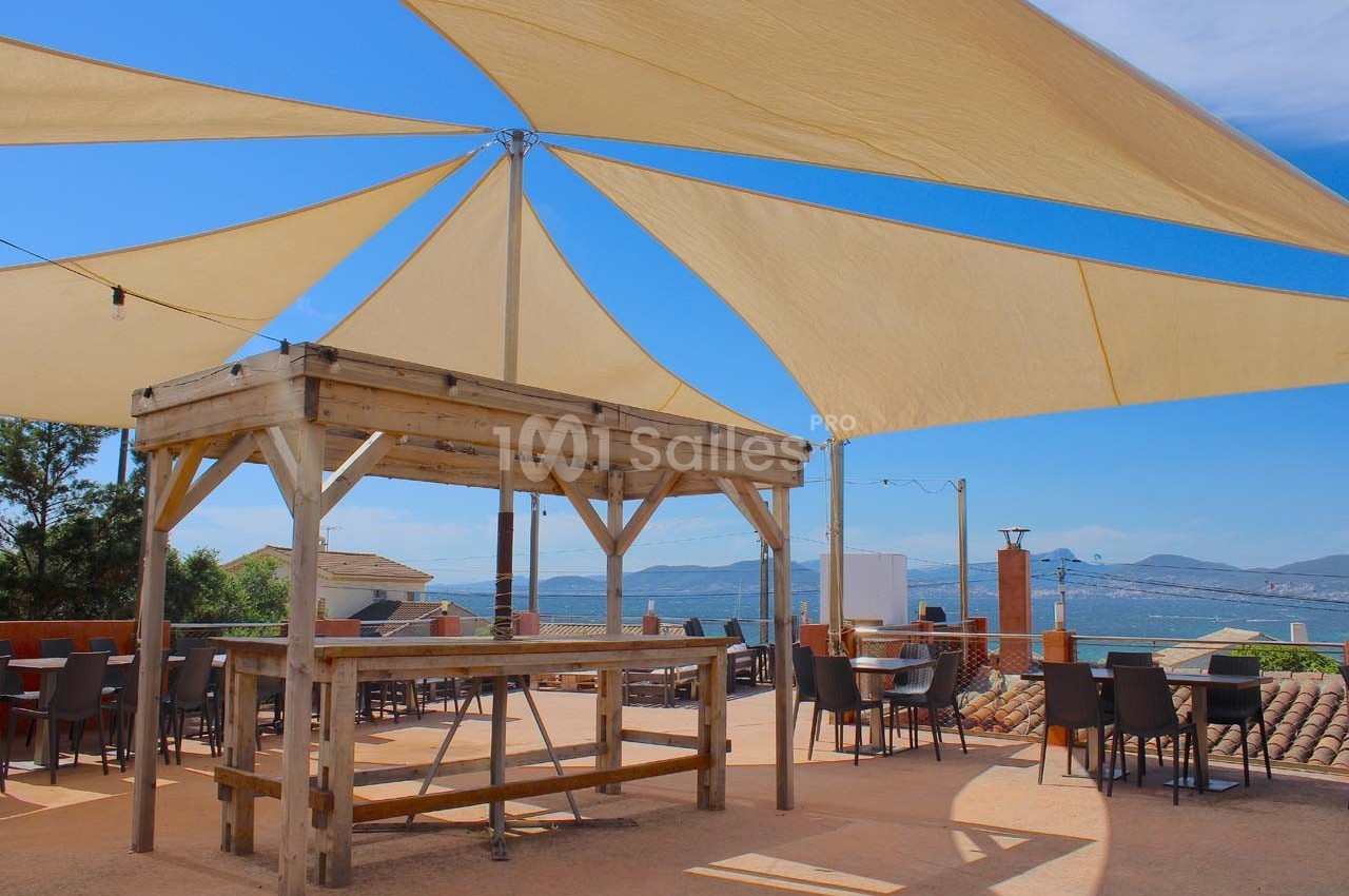 Terrasse ensoleillée avec voiles d'ombrage, tables et chaises, offrant une vue sur la mer et les collines au loin.