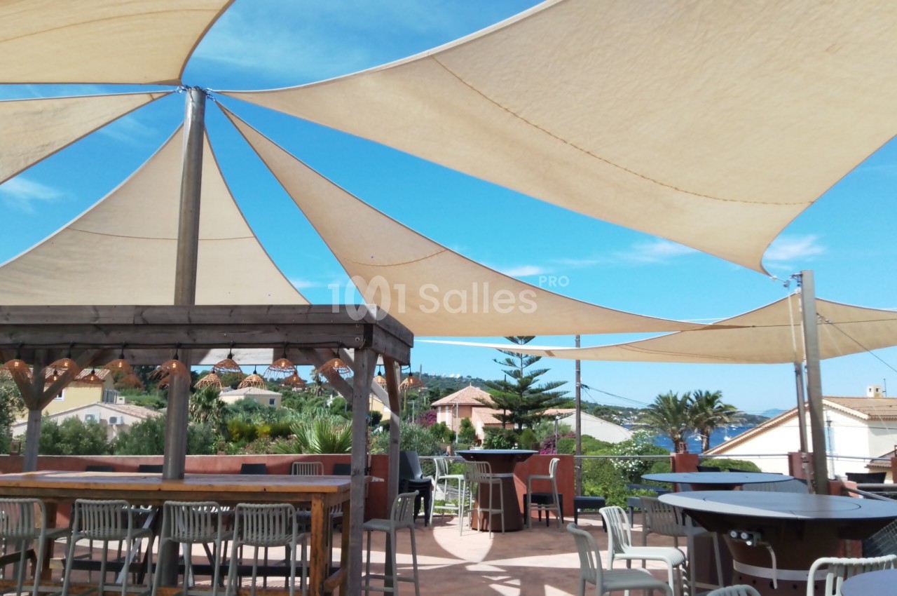 Terrasse ensoleillée avec voiles d'ombrage, tables et chaises, entourée de végétation et maisons en arrière-plan.
