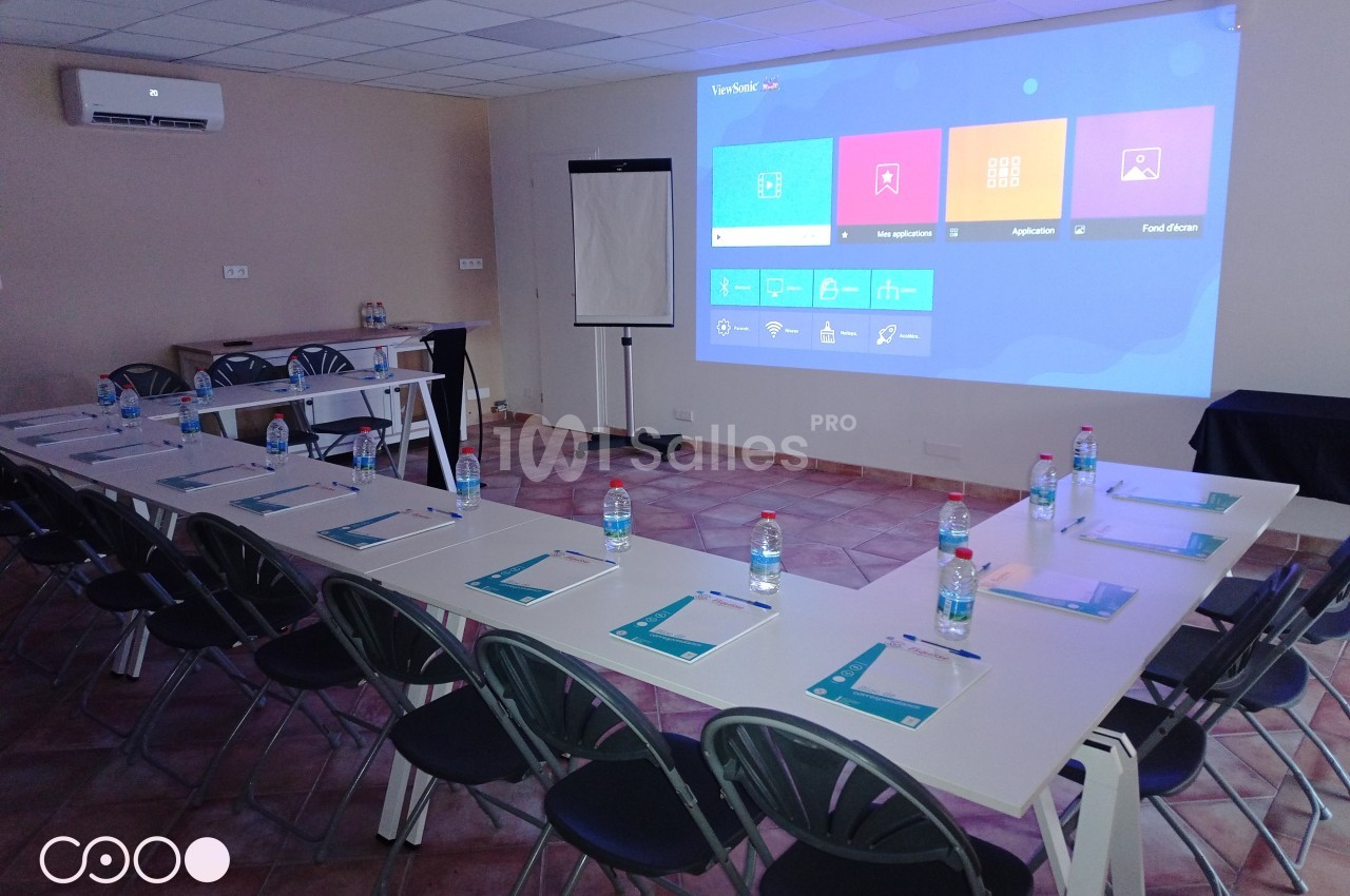 Salle de réunion équipée avec tables en U, chaises, écran de projection, paperboard et documents disposés sur les tables.