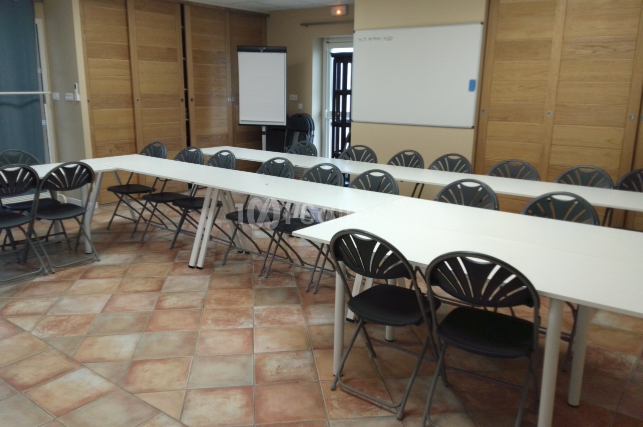Salle de réunion avec des tables blanches disposées en U, des chaises noires et un tableau blanc au mur.