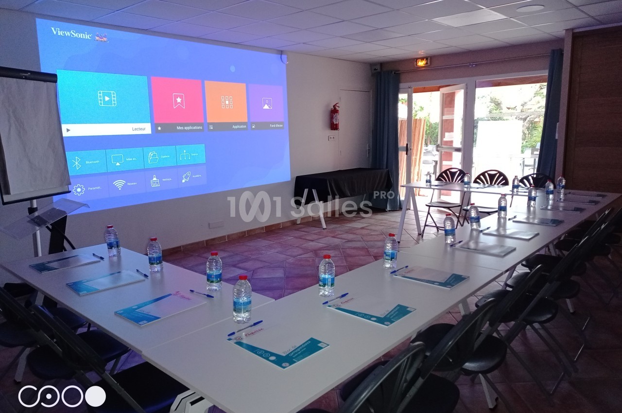 Salle de réunion équipée d'un projecteur affichant un écran d'accueil, tables disposées en U avec documents et bouteilles d…