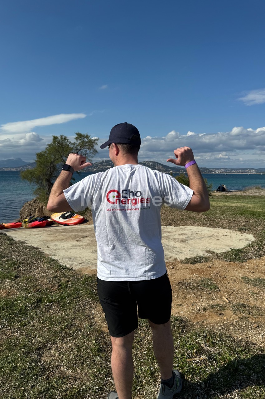 Un homme de dos portant un t-shirt blanc avec un logo, debout sur une pelouse près d'un lac par temps ensoleillé.