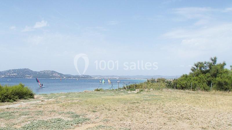 Vue d'une plage avec des planches à voile sur l'eau, des collines en arrière-plan et de la végétation au premier plan.