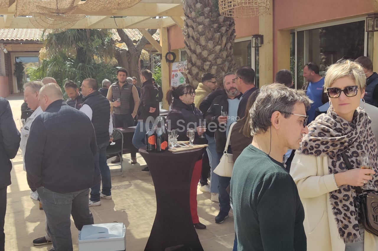 Un groupe de personnes discute en extérieur sous une pergola, avec des tables et des décorations en osier.