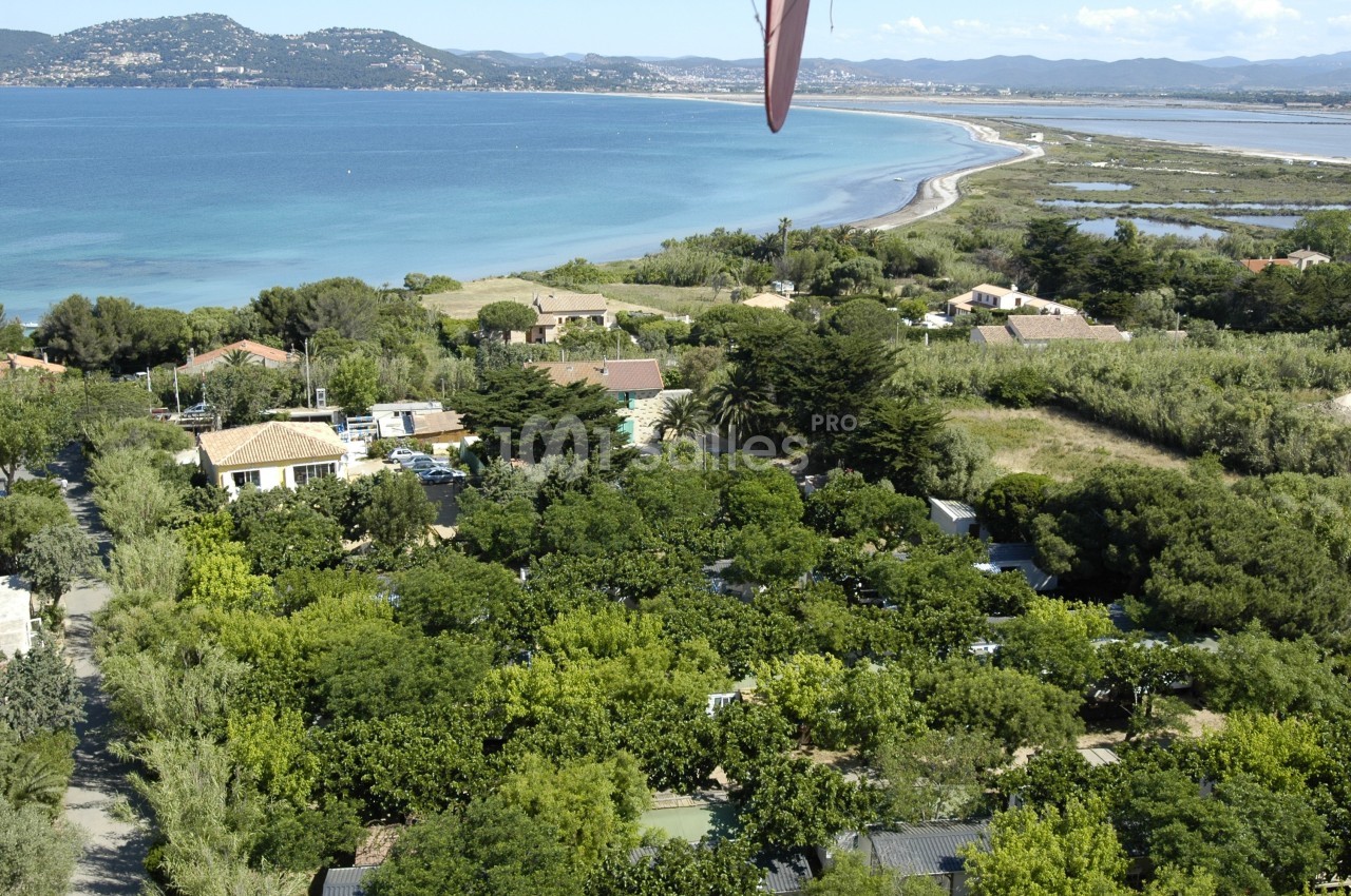 Vue aérienne d'un paysage côtier avec des maisons entourées de végétation dense, une plage et la mer en arrière-plan.
