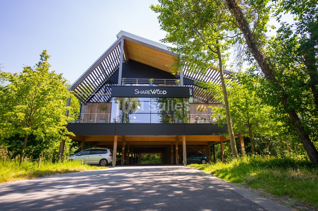 Bâtiment moderne sur pilotis entouré d'arbres, avec une façade vitrée et un panneau ’Sharewood’.