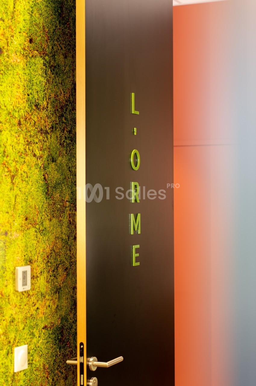 Porte noire entrouverte avec l'inscription ’L'ORME’ en lettres vertes, mur végétal en arrière-plan.