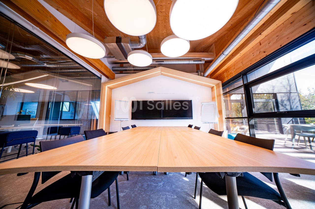 Salle de réunion moderne avec une grande table en bois, des chaises noires, des écrans et un éclairage suspendu.