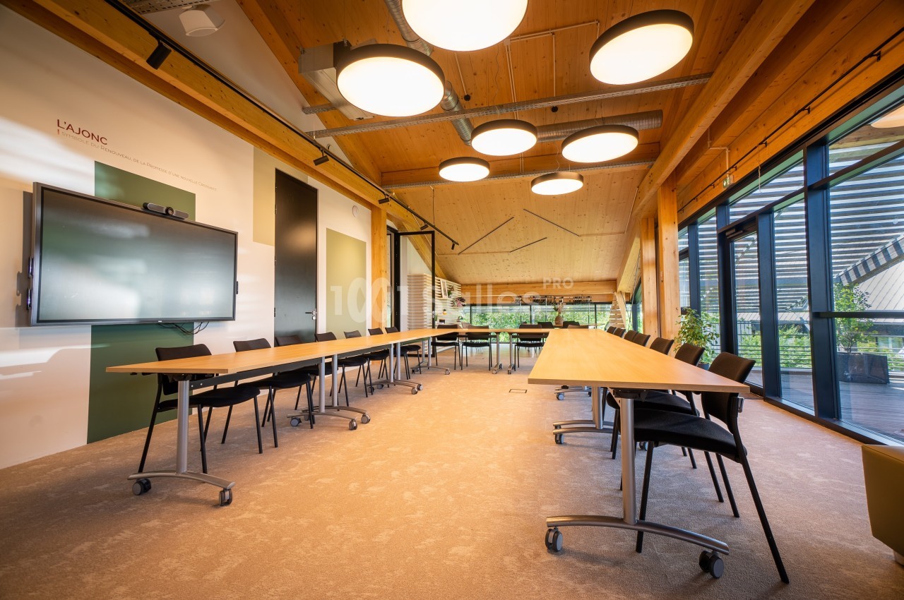 Salle de réunion lumineuse avec grandes tables en bois, chaises noires, écran interactif et larges baies vitrées.