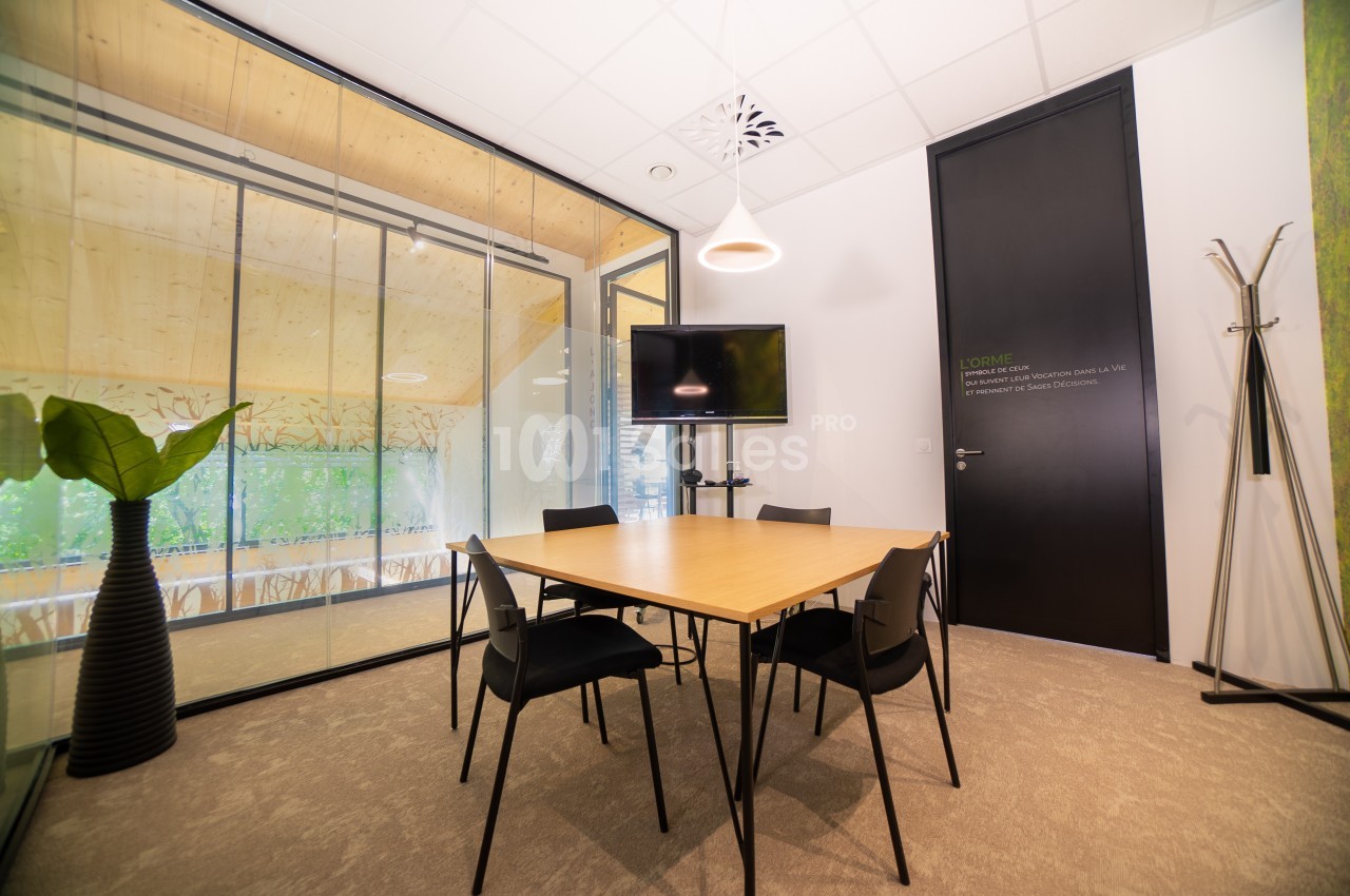 Salle de réunion lumineuse avec table en bois, quatre chaises noires, écran mural et cloison vitrée donnant sur l'extérieur.