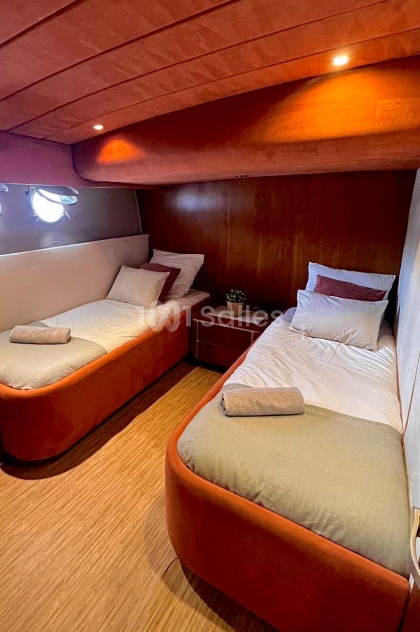 Cabine de bateau avec deux lits simples, linge de lit beige et rouge, éclairage intégré et hublots.