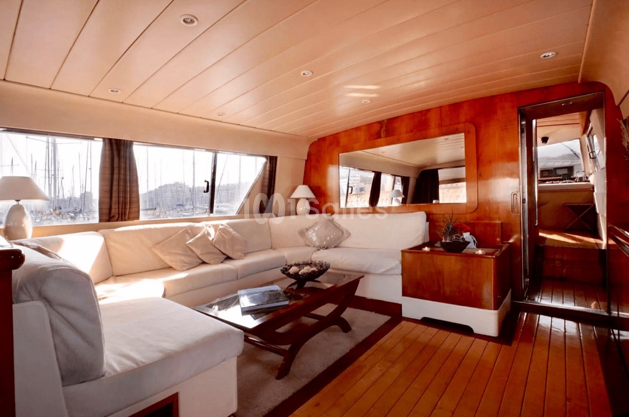 Intérieur d'un yacht avec salon spacieux, canapé blanc, table basse en bois et grandes fenêtres offrant une vue sur le port.