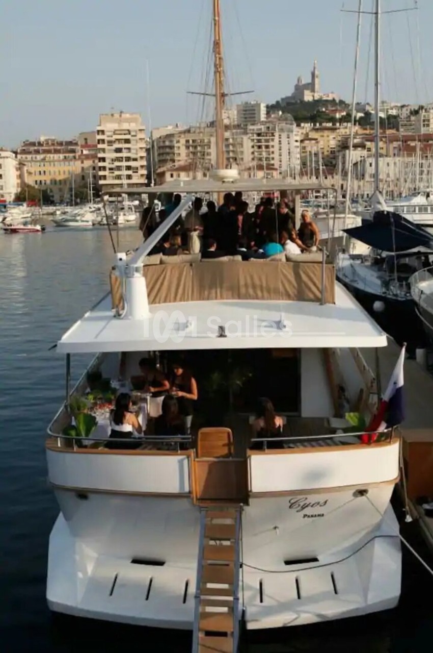 Vue arrière d'un yacht amarré dans un port, avec des personnes rassemblées sur le pont supérieur.