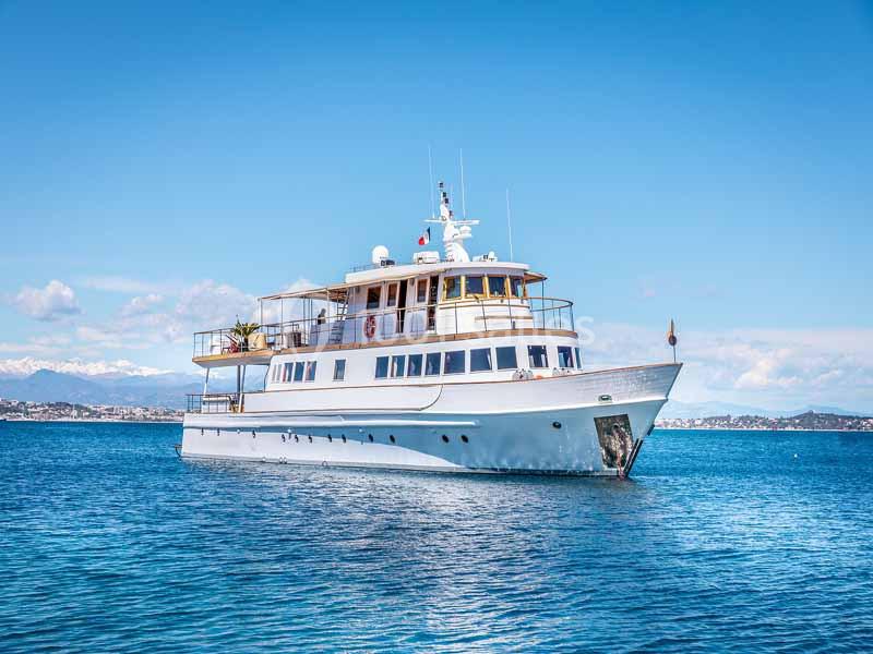 Yacht blanc naviguant sur une mer calme sous un ciel bleu, avec une côte visible à l'arrière-plan.