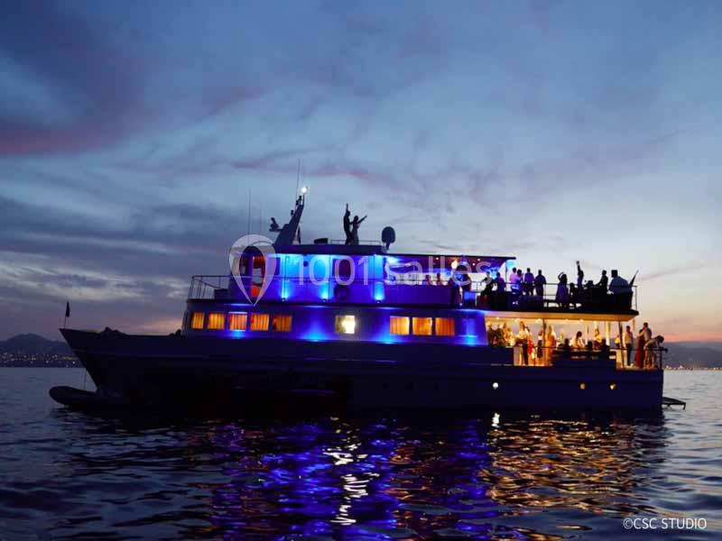 Bateau éclairé en bleu naviguant au crépuscule avec des passagers visibles sur le pont.