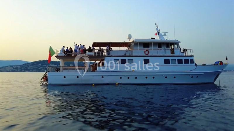 Un grand yacht blanc navigue sur une mer calme au coucher du soleil, avec des passagers sur le pont supérieur.
