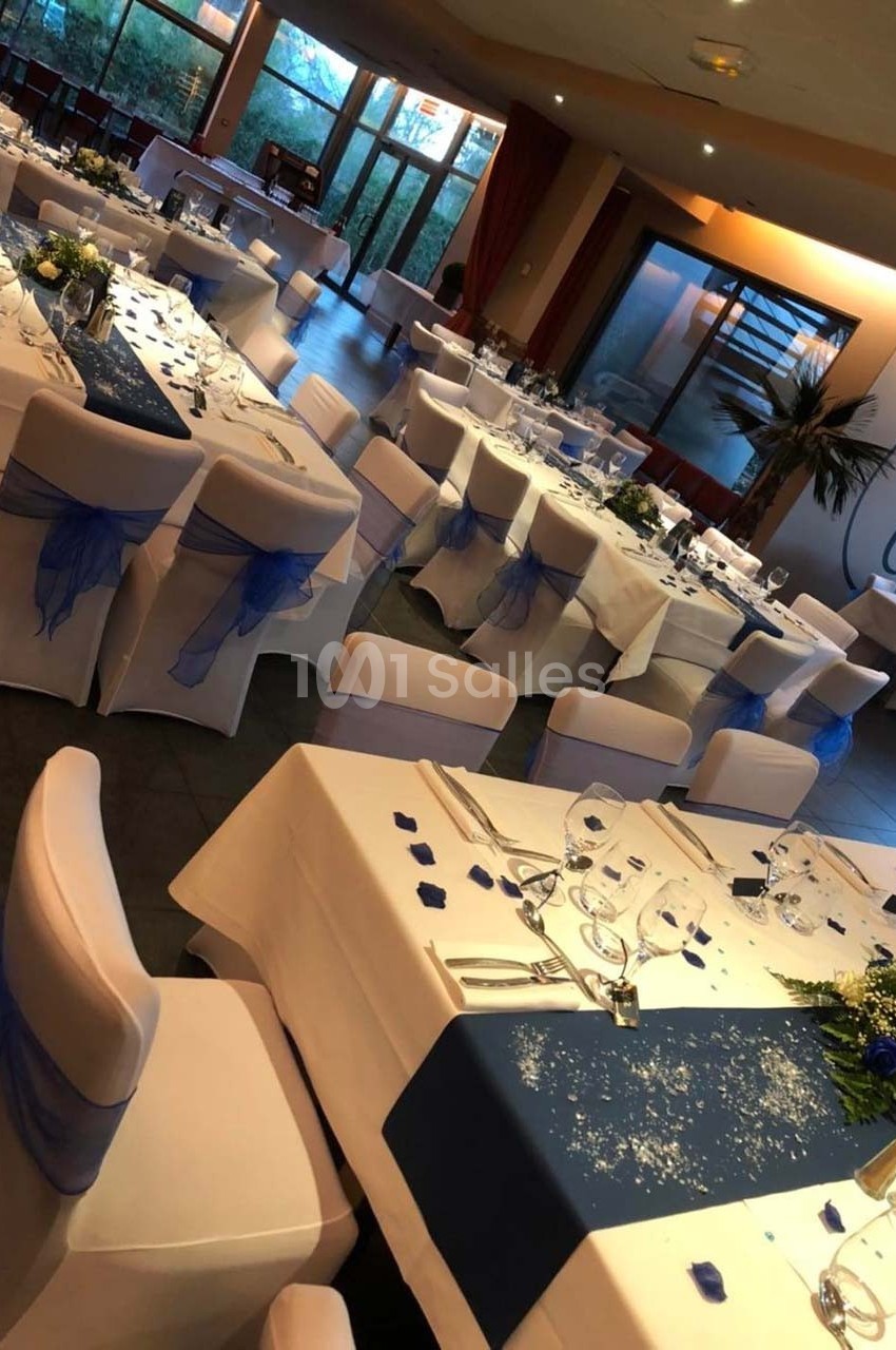 Salle de réception décorée avec des tables dressées, nappes blanches et chemins de table bleus, ambiance lumineuse.