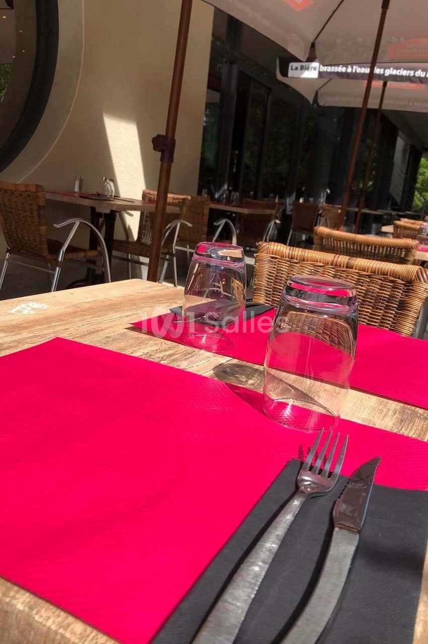 Terrasse de restaurant avec tables en bois, nappes rouges, couverts et verres, entourée de chaises en osier.