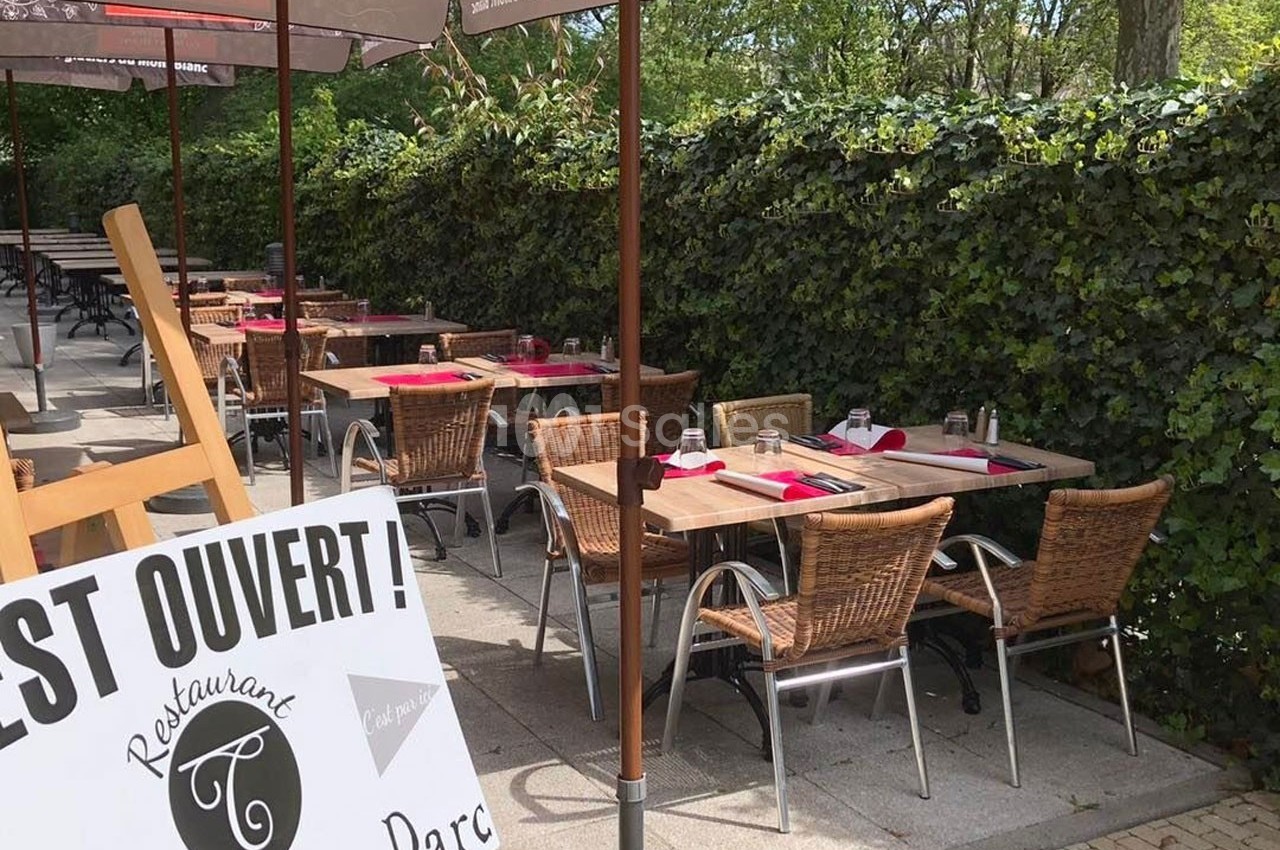 Terrasse d'un restaurant avec des tables dressées et des chaises en rotin, entourée de verdure et protégée par des parasols.
