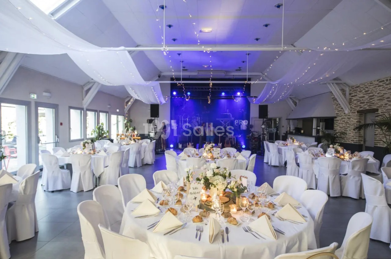 Salle de réception décorée pour un événement, avec tables dressées et scène éclairée en arrière-plan.
