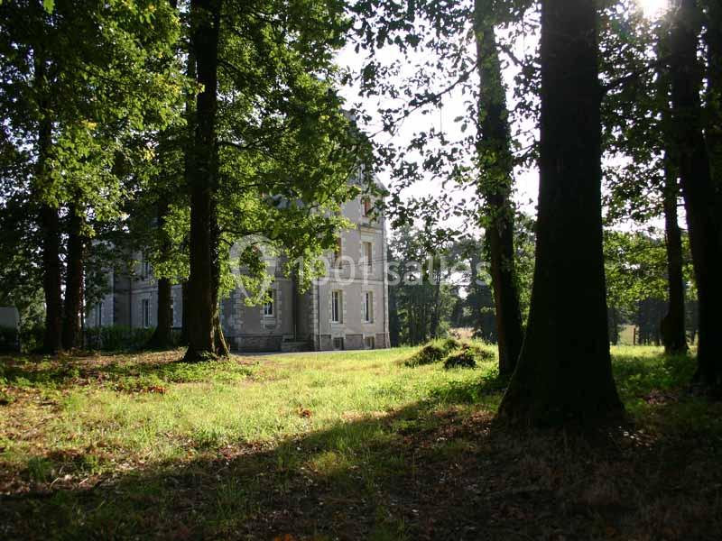 Manoir en pierre partiellement ombragé, entouré d'arbres dans un parc verdoyant baigné de lumière naturelle.