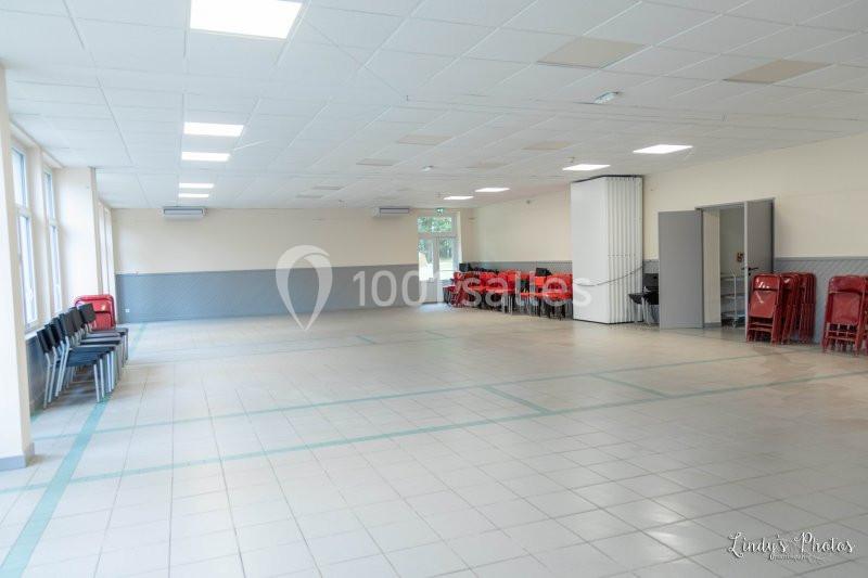 Salle vide avec carrelage clair, chaises alignées contre les murs et empilées dans un coin, lumière naturelle par des…