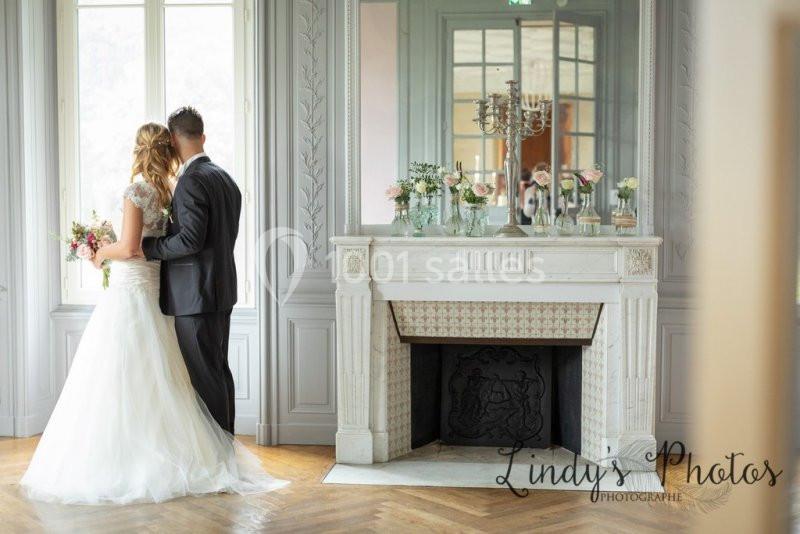 Un couple en tenue de mariage se tient près d'une fenêtre dans une pièce élégante avec une cheminée décorée.