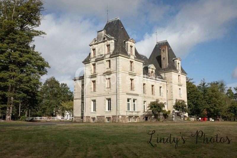 Château ancien en pierre avec toit mansardé, entouré d'arbres et situé sur une pelouse sous un ciel partiellement nuageux.
