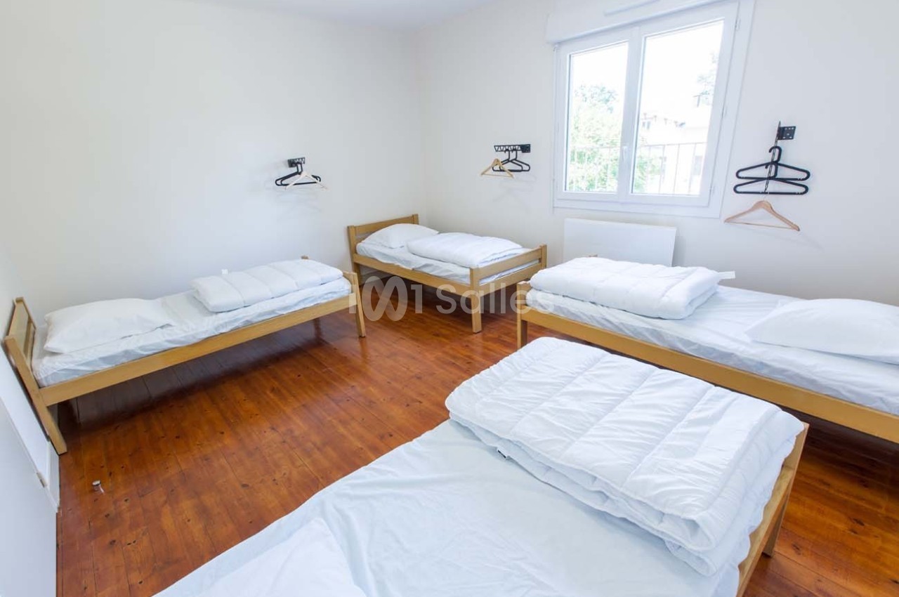Chambre lumineuse avec quatre lits simples, draps blancs, parquet en bois et cintres accrochés au mur.
