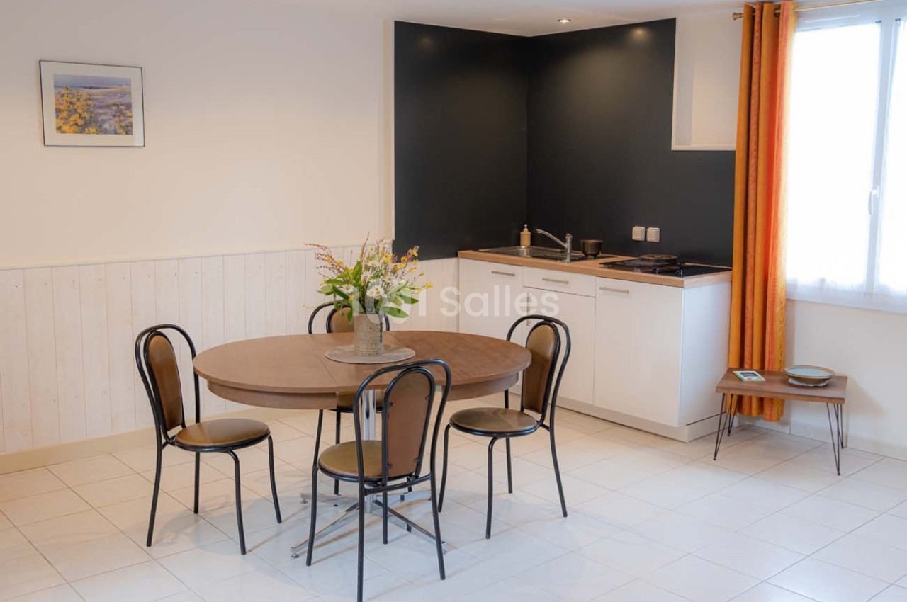 Salle à manger avec table ronde, quatre chaises, une kitchenette au fond et une fenêtre avec rideaux orange.
