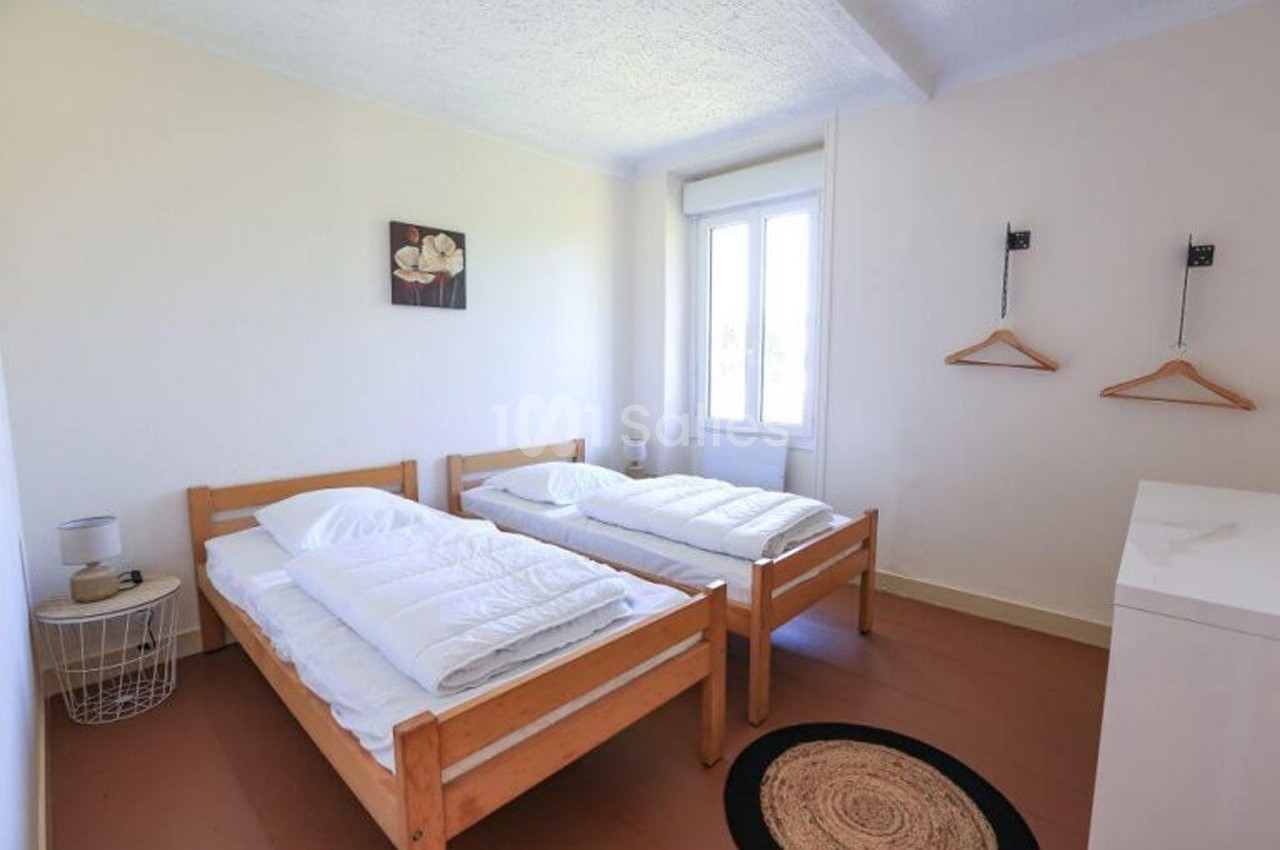 Chambre lumineuse avec deux lits simples en bois, murs blancs, fenêtre, table de chevet et cintres accrochés au mur.