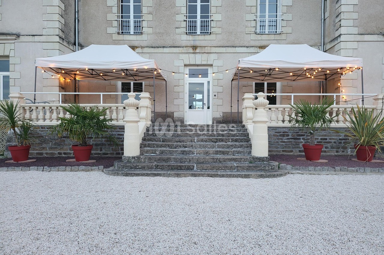 Entrée d'un bâtiment ancien avec escaliers en pierre, deux tonnelles blanches et des plantes en pots rouges.