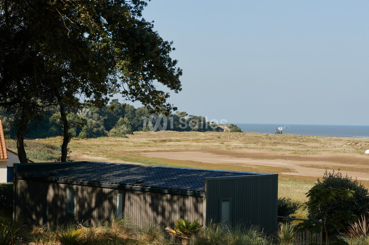 Vue d'une maison moderne entourée de végétation, avec un paysage côtier et la mer en arrière-plan.