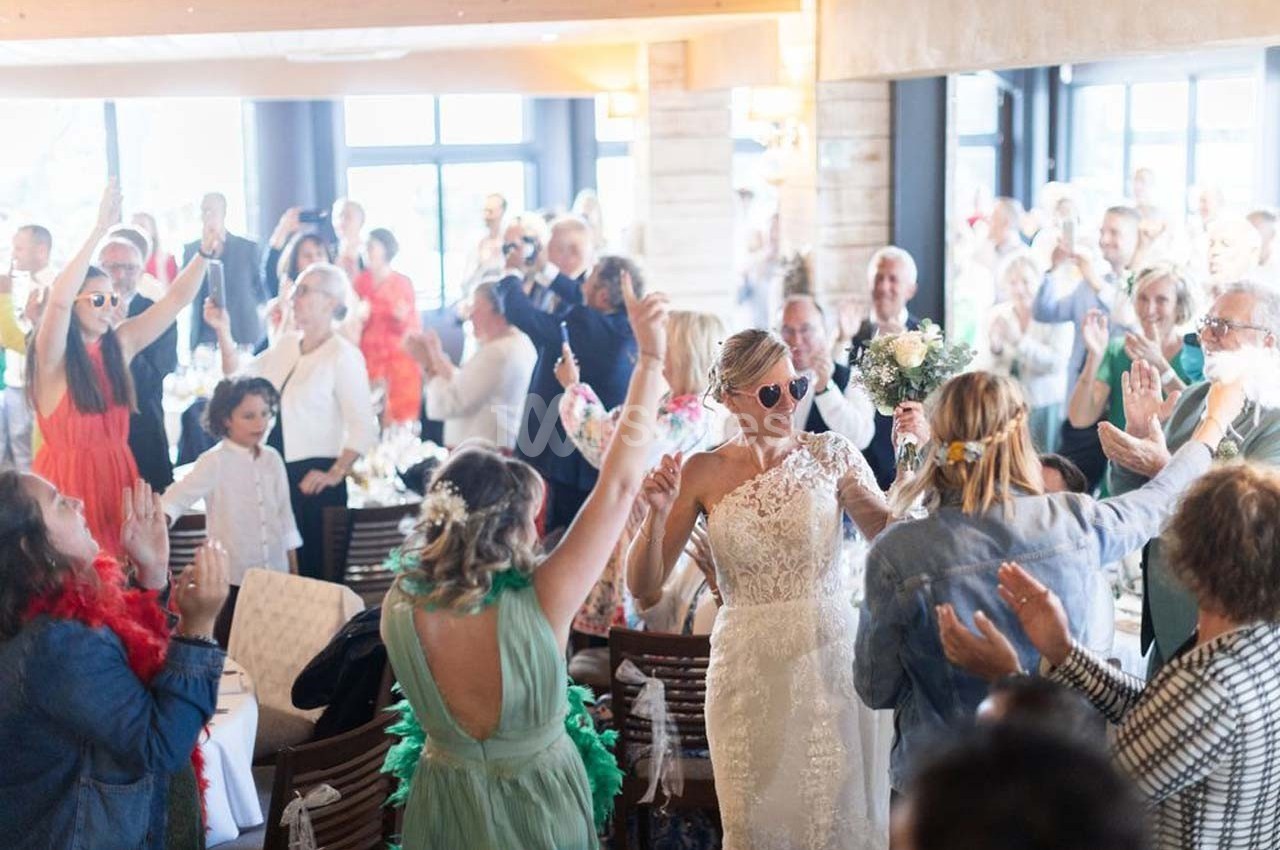 Une mariée en robe blanche entre dans une salle festive, entourée d'invités applaudissant et célébrant.