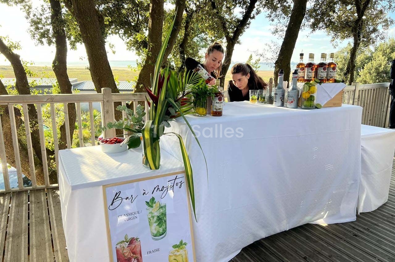 Deux personnes préparent des cocktails derrière un bar extérieur décoré, avec des bouteilles et des plantes en arrière-plan.