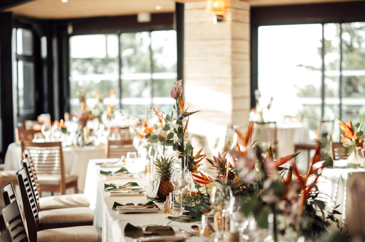Salle lumineuse avec tables élégamment dressées, décorées de fleurs tropicales et entourées de chaises en bois.