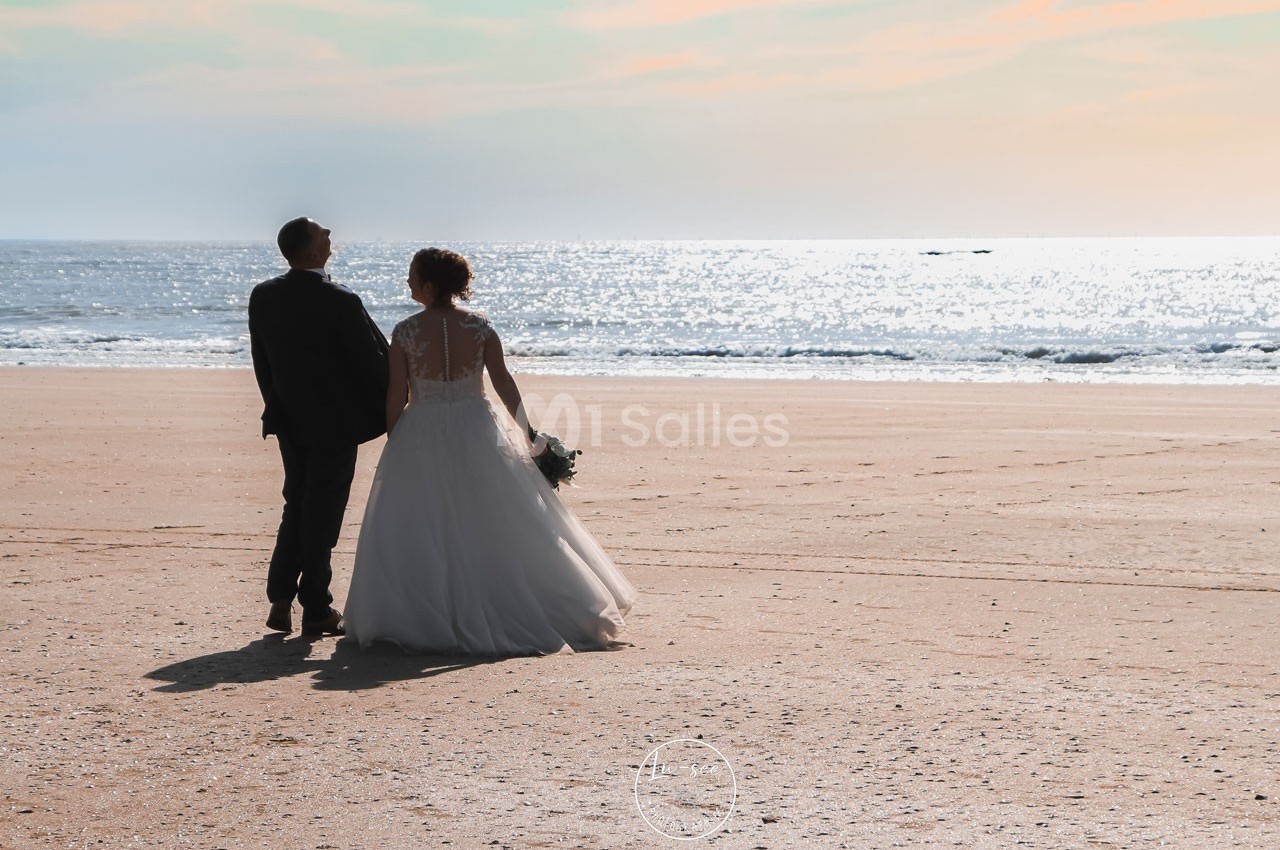 Un couple en tenue de mariage marche sur une plage ensoleillée, face à la mer scintillante.