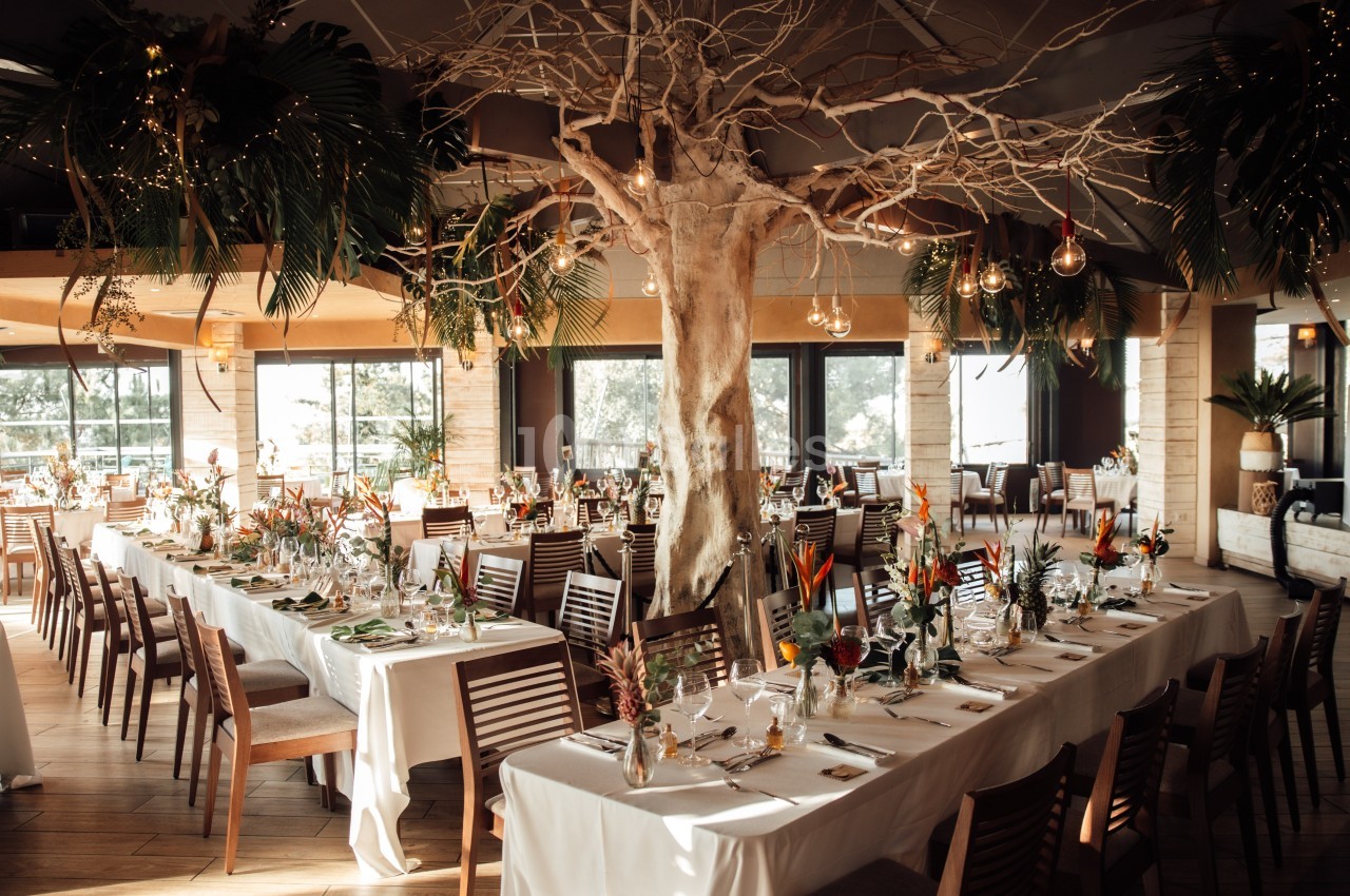 Salle de réception lumineuse avec de longues tables décorées de fleurs tropicales et un arbre central.