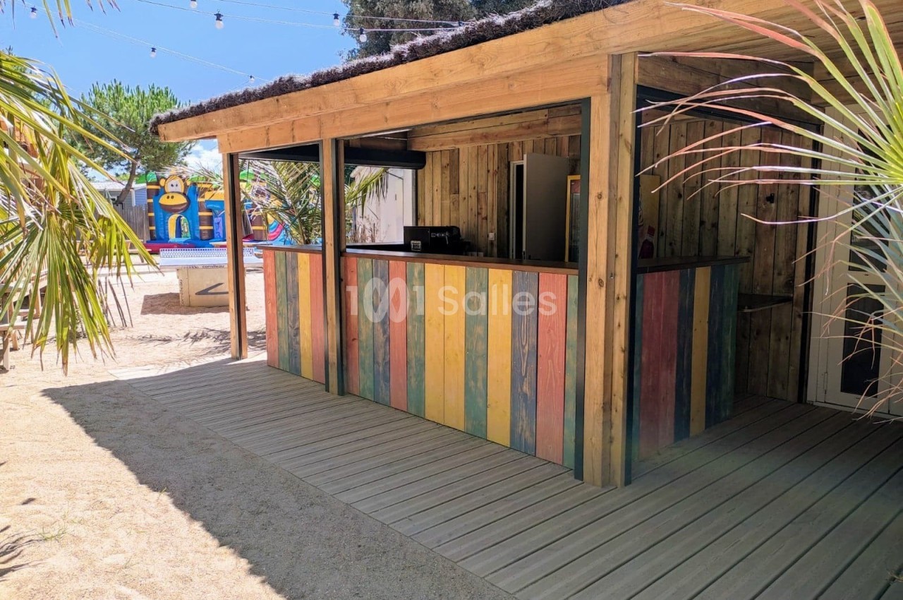 Kiosque en bois avec comptoir coloré, situé dans un espace extérieur entouré de palmiers et de décorations estivales.