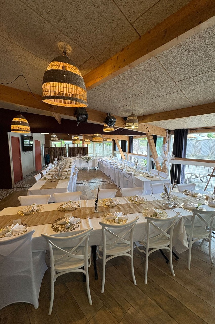 Salle de réception décorée avec de longues tables dressées, nappes blanches et éclairage suspendu en osier.