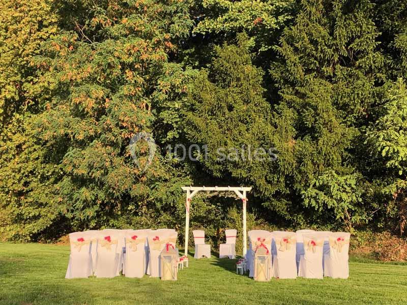 Chaises décorées et arche blanche installées en extérieur pour une cérémonie, devant un fond d'arbres verdoyants.