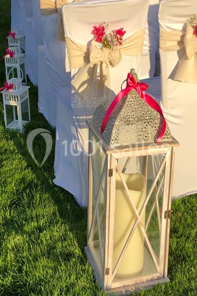 Grande lanterne blanche avec bougie, posée sur de l'herbe, entourée de chaises décorées de rubans et fleurs roses.