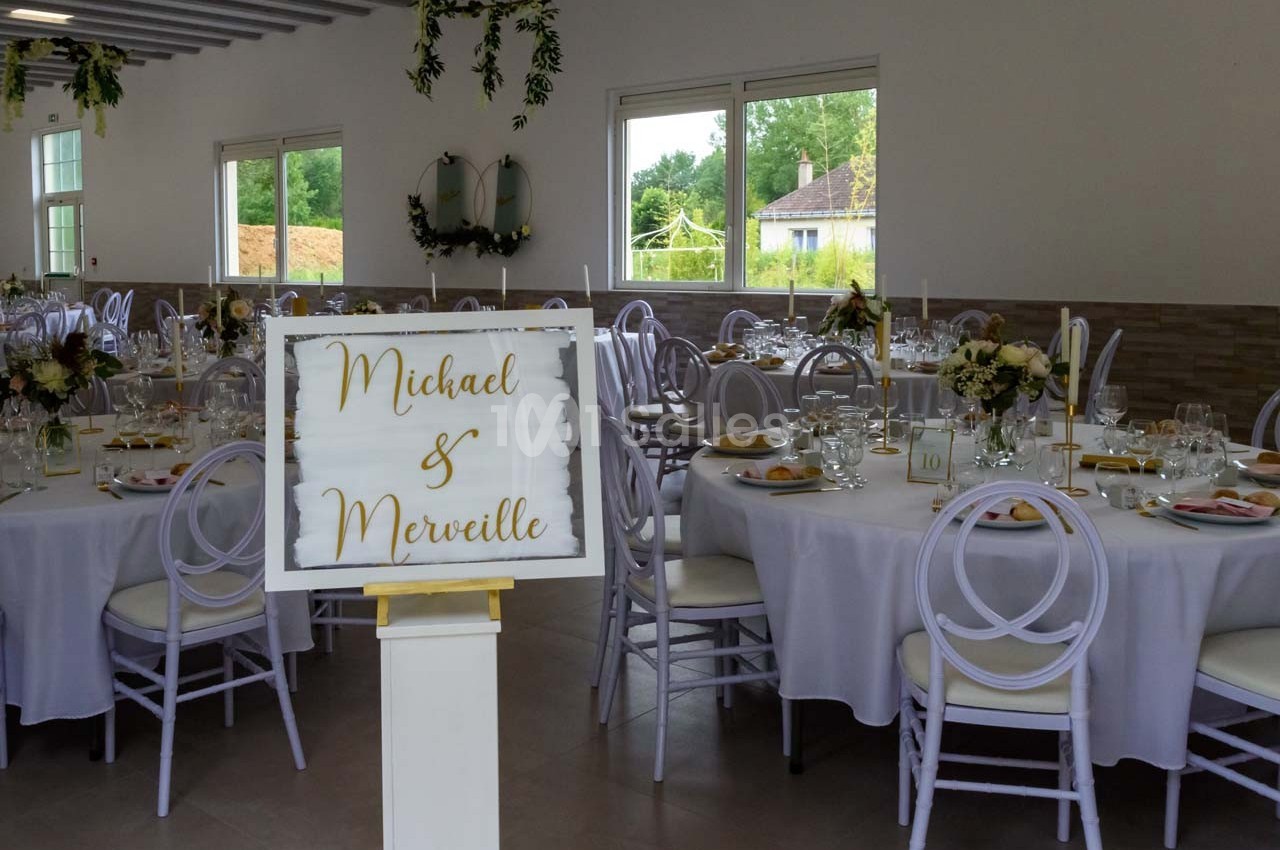 Salle de réception décorée pour un mariage, avec des tables dressées et un panneau portant des prénoms.
