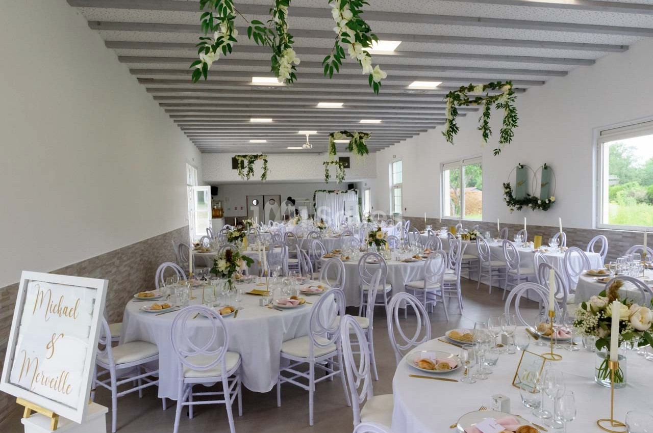 Salle de réception décorée pour un mariage, avec tables rondes, chaises blanches et ornements floraux suspendus.